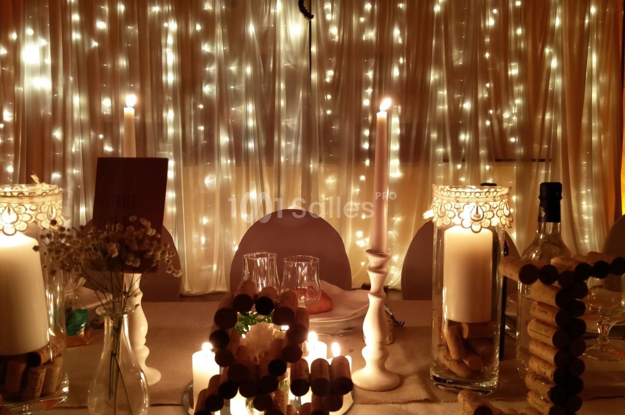 Table décorée avec des bougies, des guirlandes lumineuses et des éléments en liège, devant un rideau éclairé.