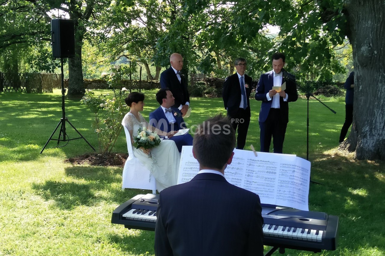 Un mariage en extérieur avec un couple assis, un pianiste jouant et un homme lisant un discours devant des invités.