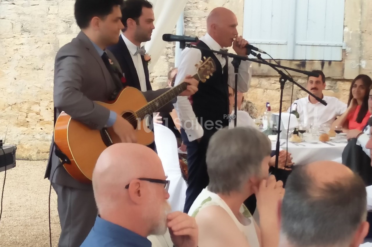 Des musiciens et un chanteur animent un repas en plein air, entourés d'invités assis à des tables.