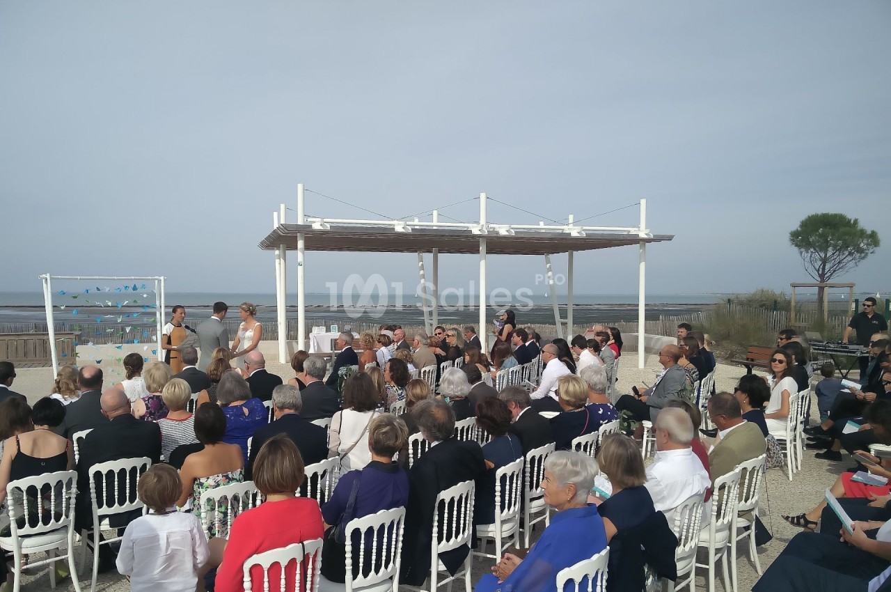 Cérémonie en plein air avec des invités assis sur des chaises blanches, face à un officiant près de la mer.