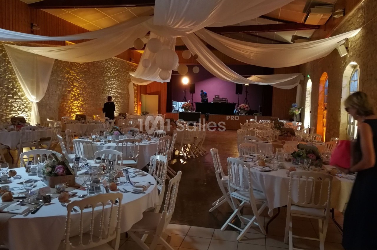 Salle de réception décorée avec des tables rondes dressées, des chaises blanches et des drapés suspendus au plafond.
