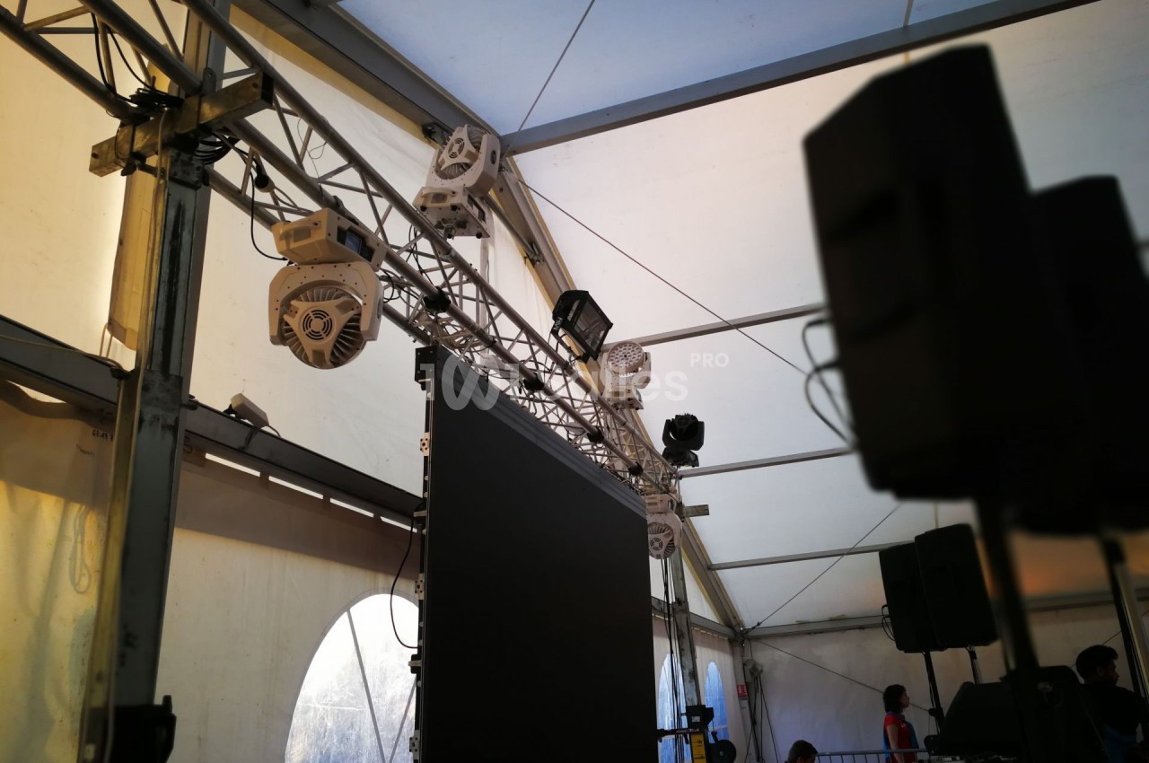 Structure métallique avec projecteurs, écran suspendu et haut-parleurs dans une tente événementielle éclairée.