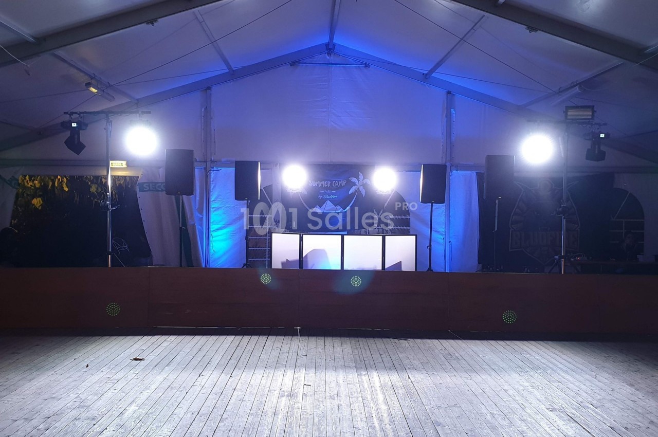 Scène sous chapiteau avec éclairage lumineux, équipement audio et piste en bois vide au premier plan.