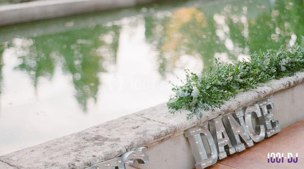 Lettres décoratives formant le mot ’DANCE’ posées sur un muret en pierre près d'un plan d'eau entouré de verdure.