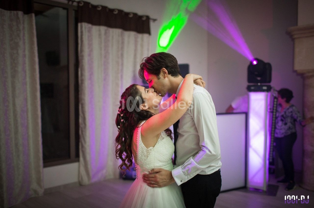 Un couple en tenue de mariage danse tendrement dans une salle éclairée par des lumières colorées.