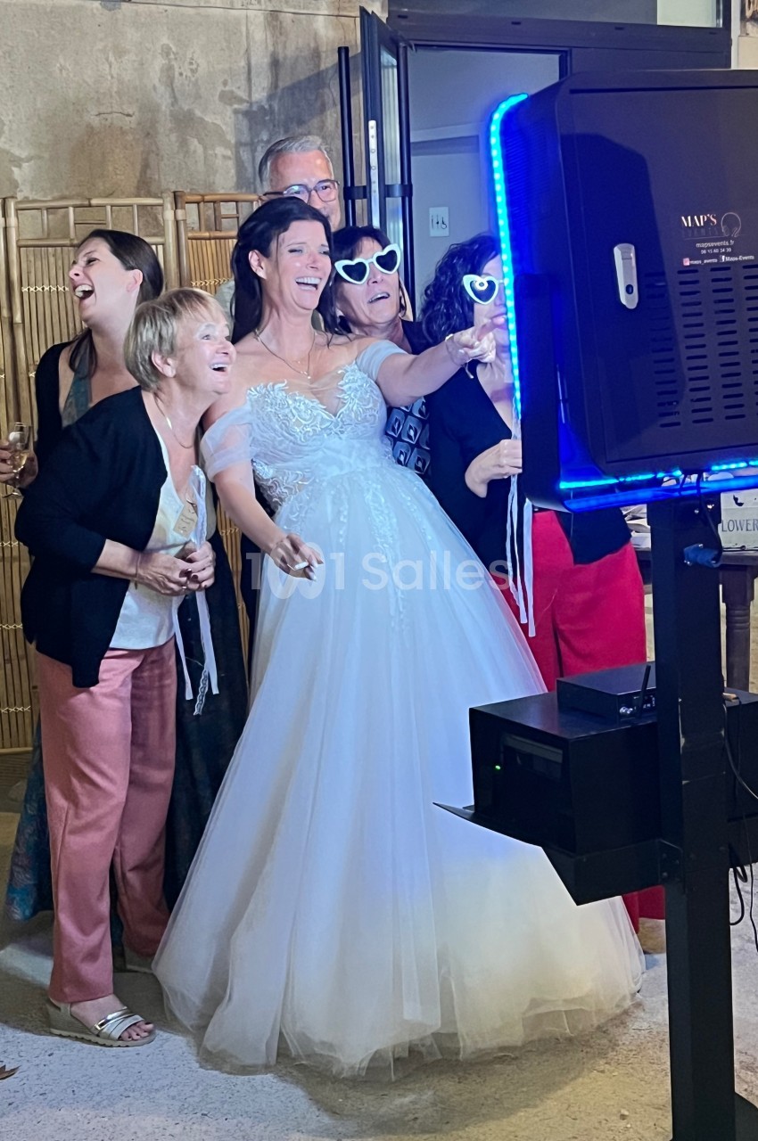 Un groupe de personnes, dont une mariée en robe blanche, pose joyeusement devant une borne photo interactive.