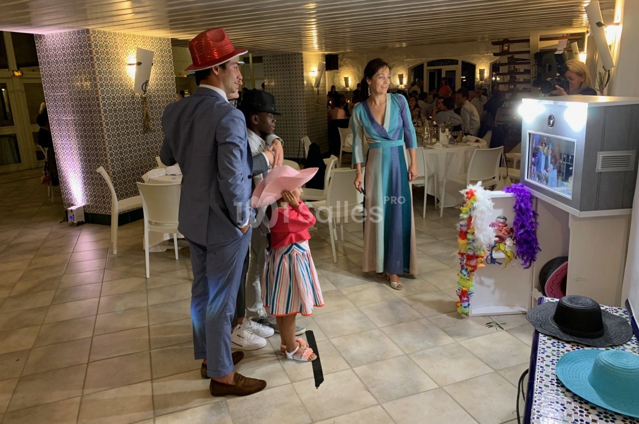 Des personnes déguisées avec des chapeaux colorés utilisent une borne photo dans une salle de réception éclairée.