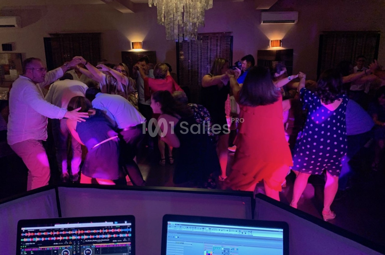 Des personnes dansent en cercle sous un éclairage coloré, vues depuis une table de mixage avec des écrans allumés.