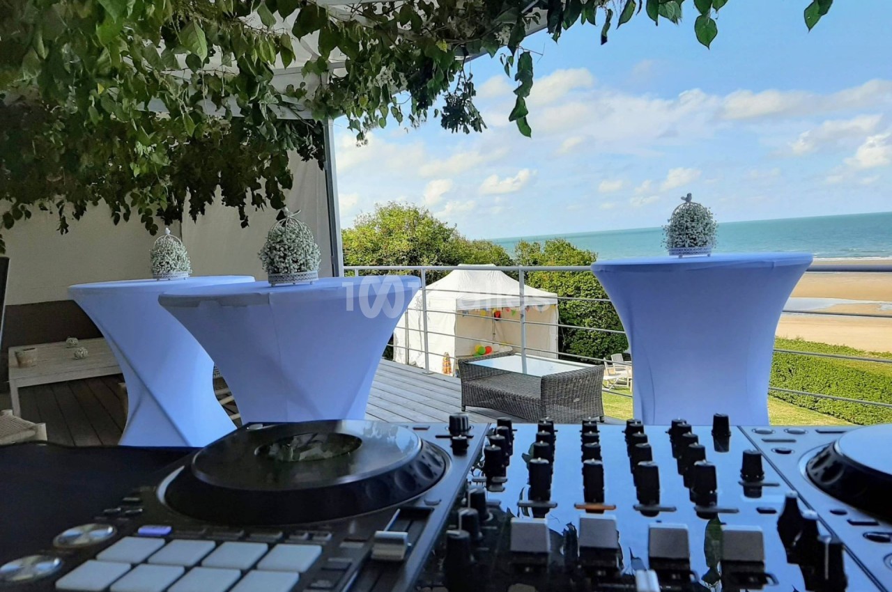 Vue d'une table de mixage en premier plan avec une terrasse décorée, donnant sur une plage et la mer en arrière-plan.