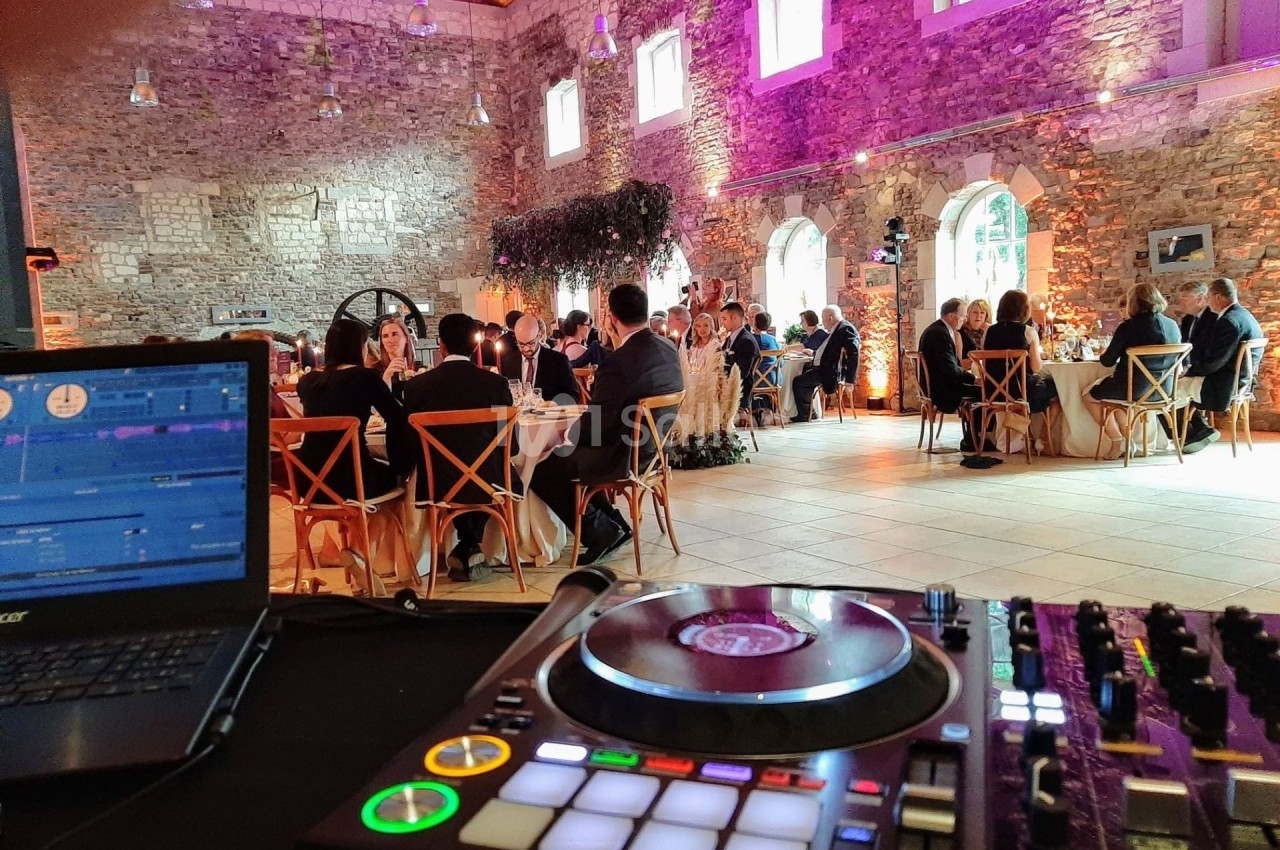 Salle de réception en pierre avec invités assis à des tables rondes, vue depuis une table de mixage de DJ au premier plan.