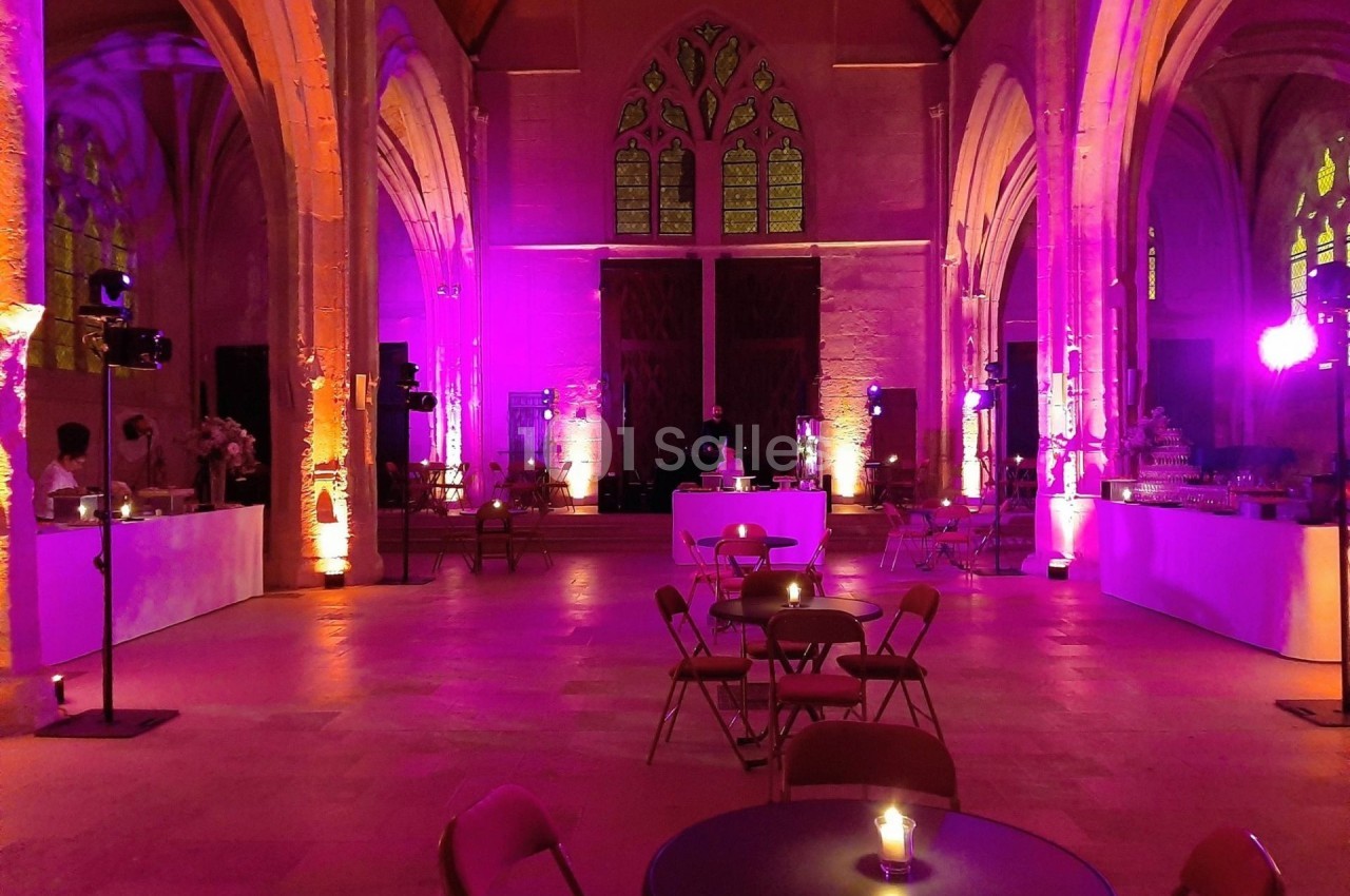 Salle voûtée éclairée de lumières violettes, avec tables et chaises disposées pour un événement.