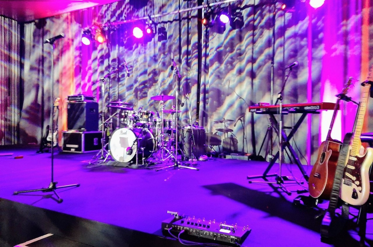 Scène de concert avec batterie, guitares, claviers et micros sous un éclairage coloré.