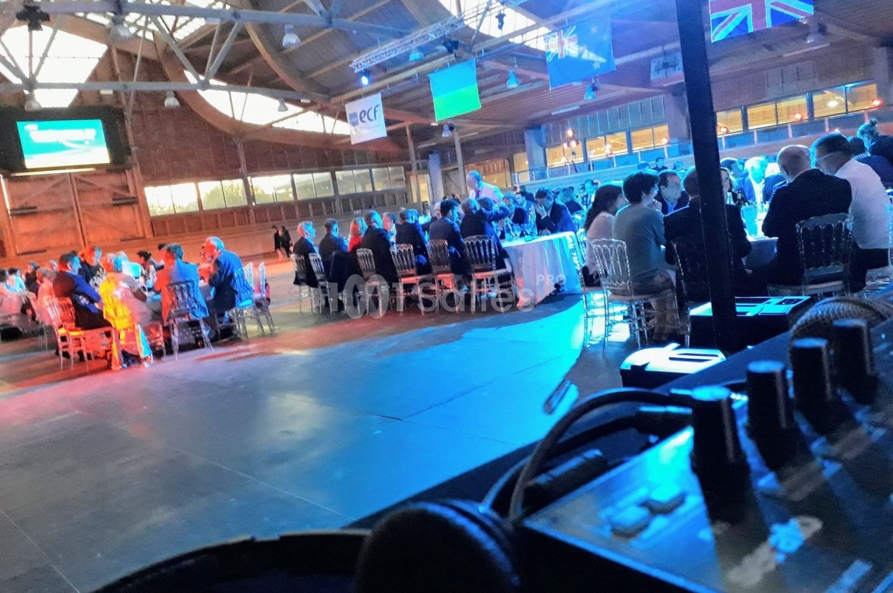 Salle de conférence avec des tables rondes occupées par des participants, éclairage bleu et drapeaux suspendus.
