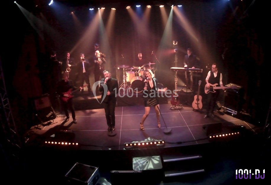 Groupe musical sur scène avec chanteurs, musiciens et éclairage coloré dans une ambiance de concert.