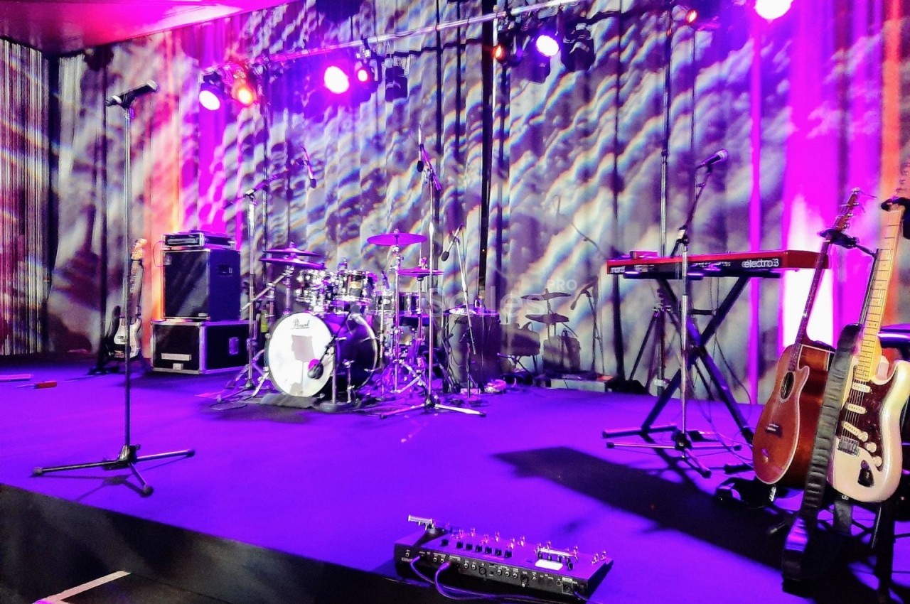 Scène de concert avec batterie, guitares, claviers et micros sous un éclairage coloré.