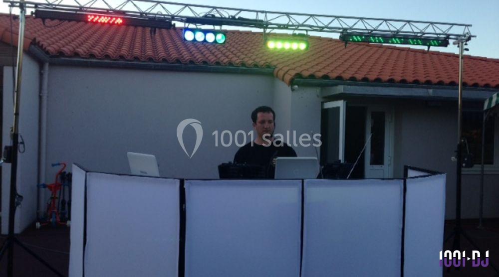 Un DJ mixe derrière une table équipée de matériel audio et éclairages colorés en extérieur, devant un bâtiment.