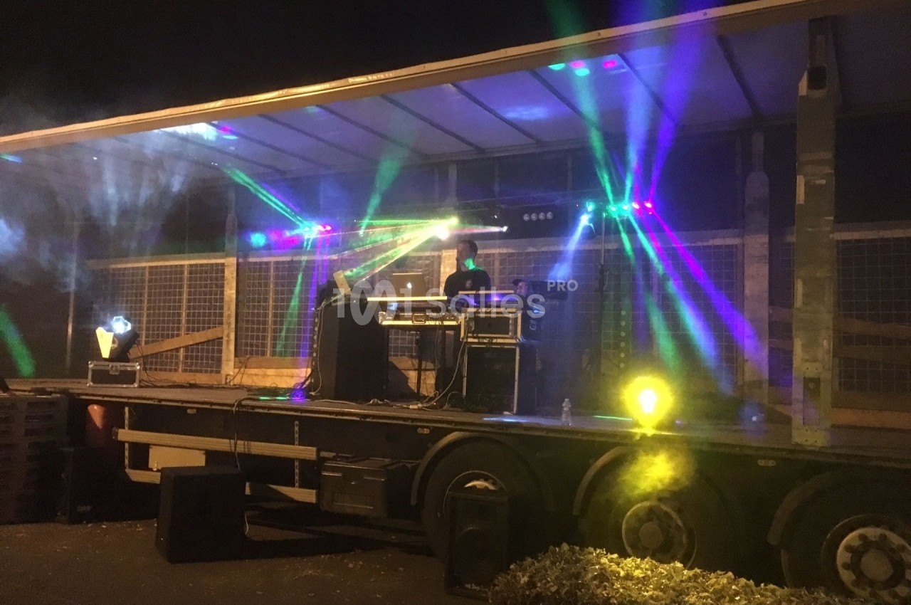 Un DJ mixe sur une scène éclairée par des jeux de lumière colorés, installée sur une remorque en extérieur.