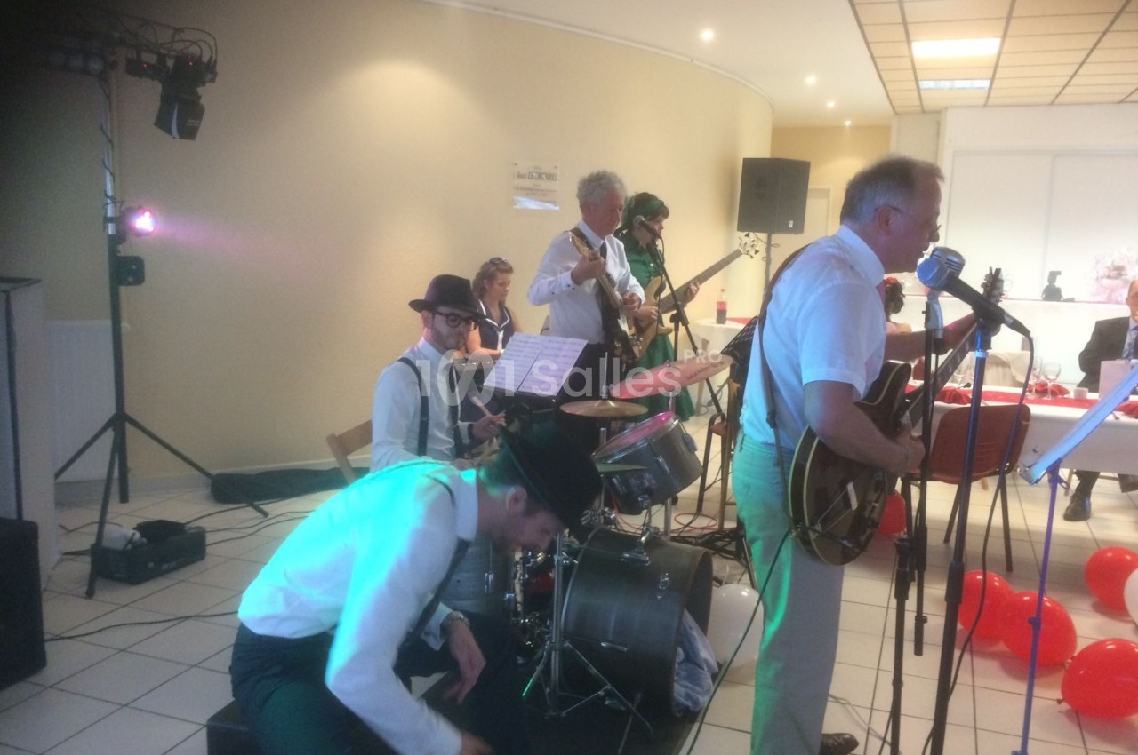 Un groupe de musiciens joue en intérieur, avec guitares, batterie et micros, devant un public hors champ.