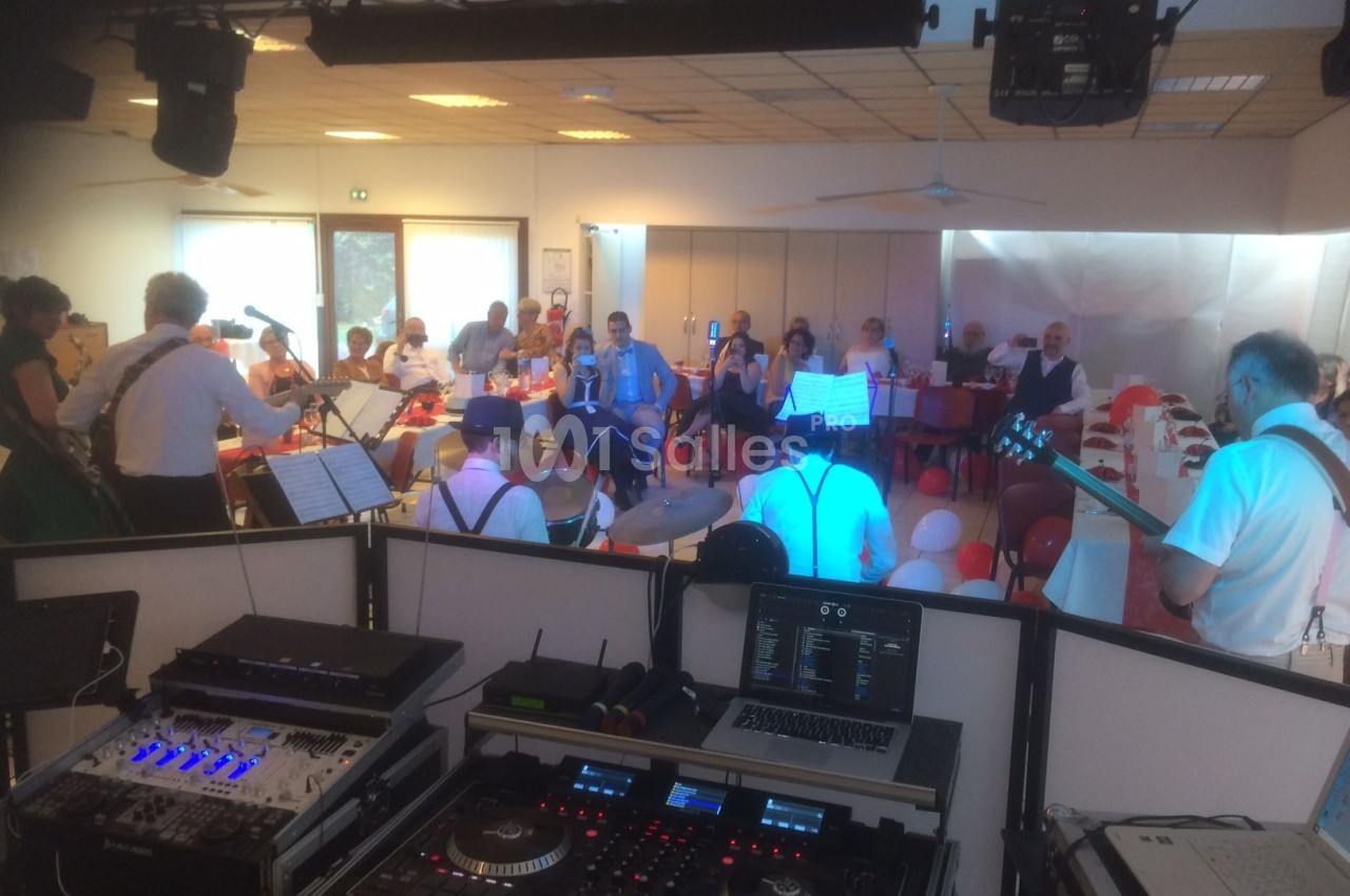 Un groupe de musiciens joue devant un public assis dans une salle éclairée, vue depuis une table de mixage.