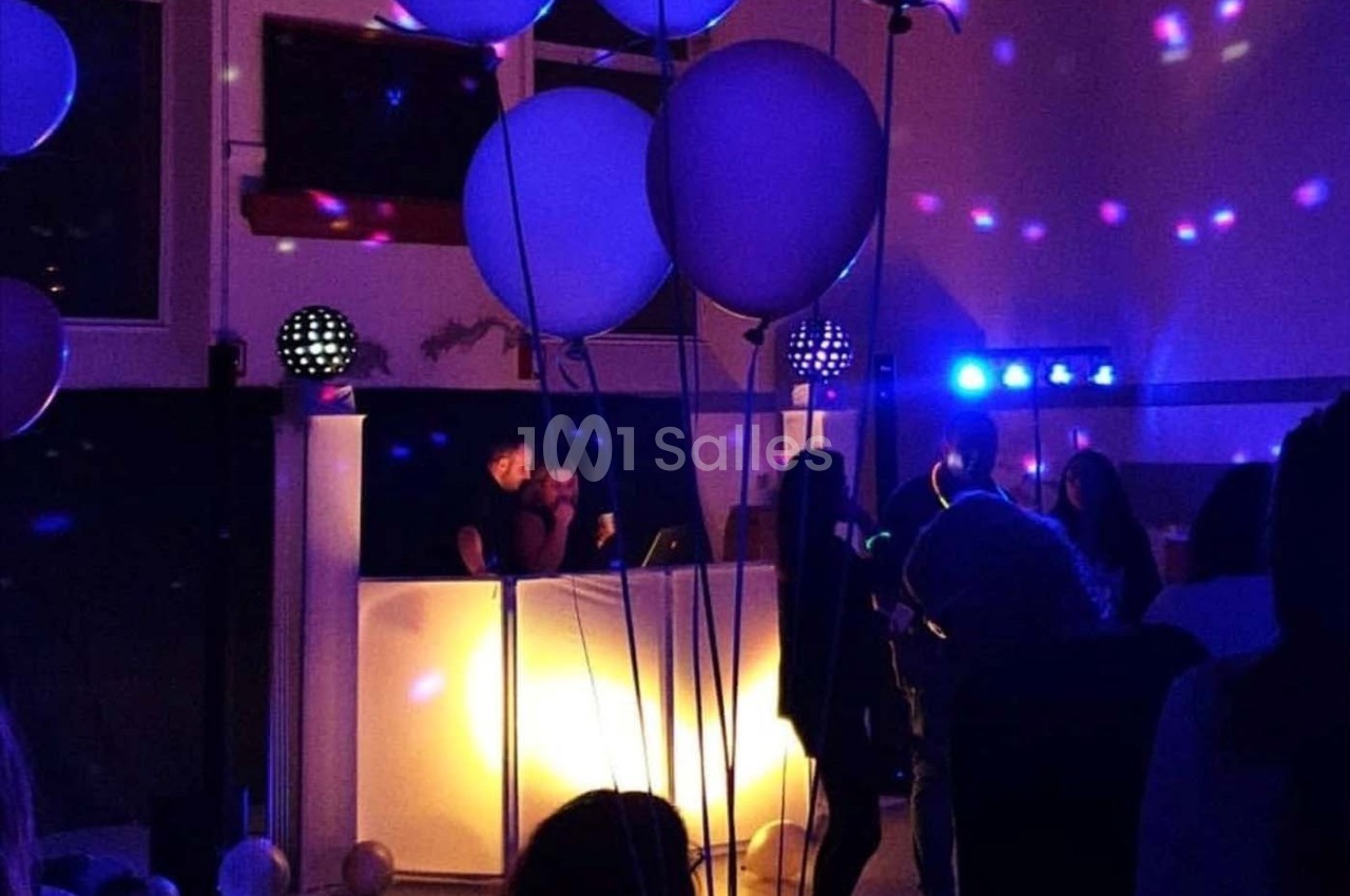 Des personnes dansent dans une salle faiblement éclairée avec un DJ en arrière-plan et des ballons suspendus.