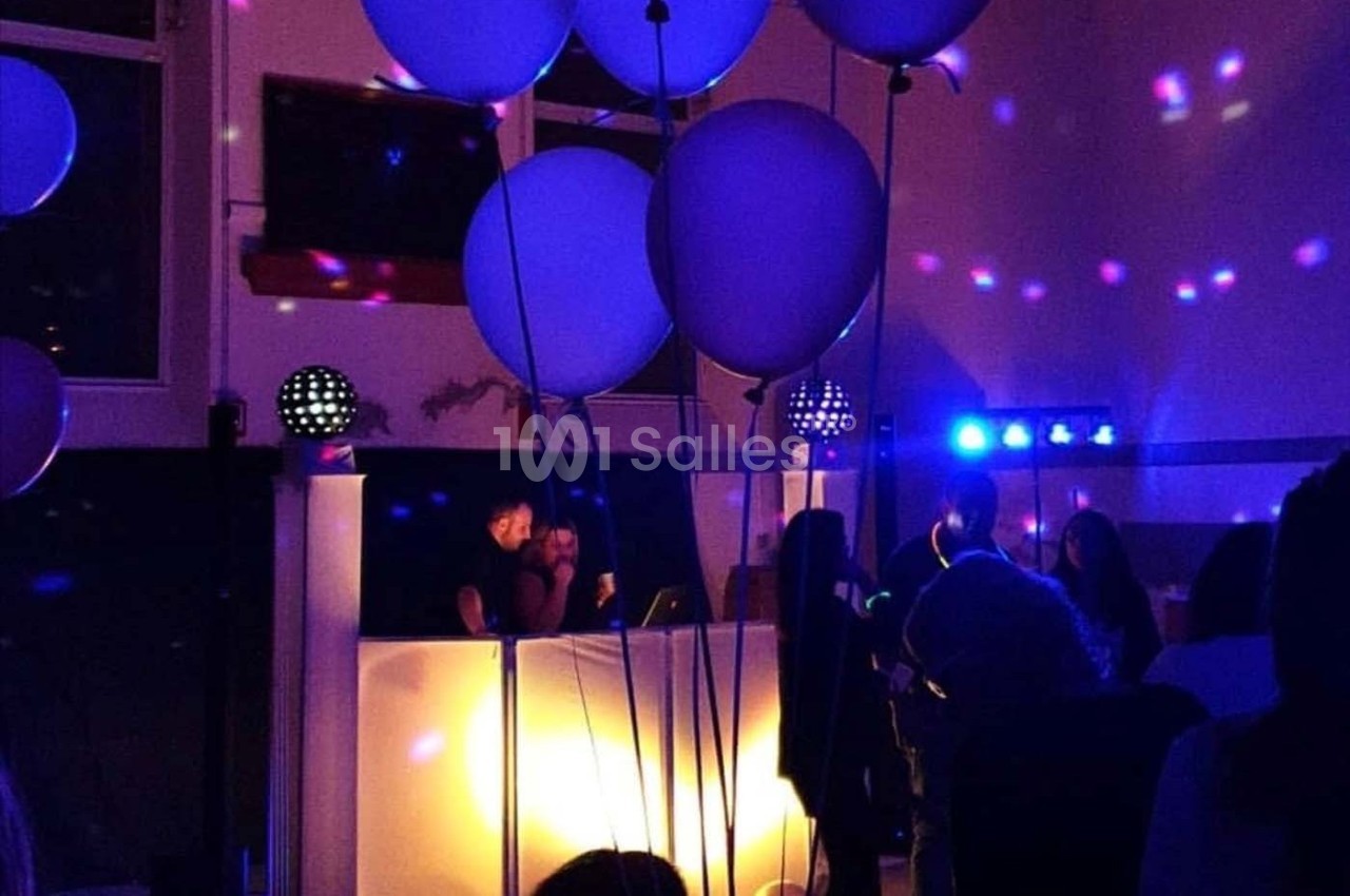 Des ballons blancs flottent dans une salle faiblement éclairée avec un DJ jouant de la musique devant un public.