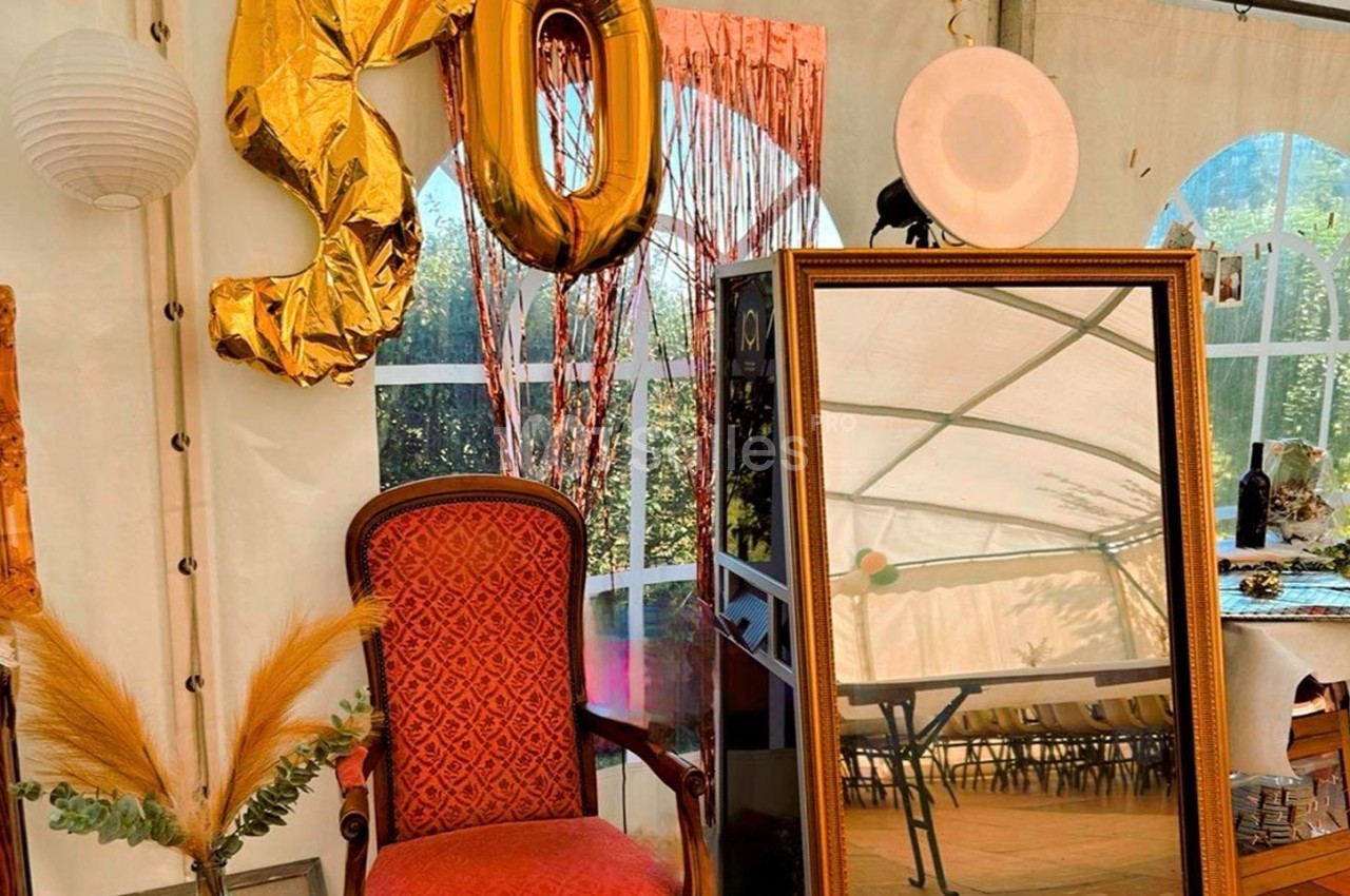 Fauteuil rouge, miroir doré, ballons ’50’ et décor festif dans une tente avec vue sur un jardin.