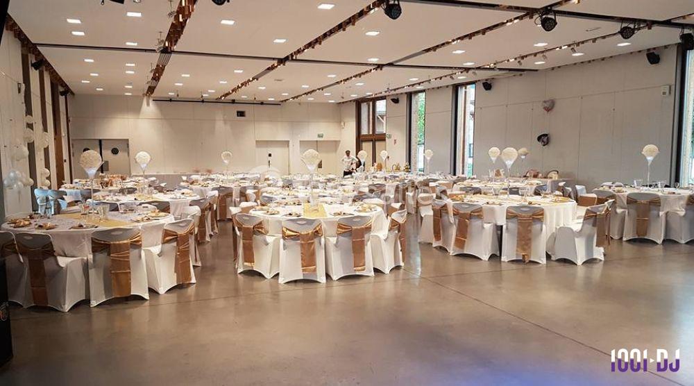 Salle de réception décorée avec des tables rondes dressées, nappes blanches et chaises ornées de rubans dorés.