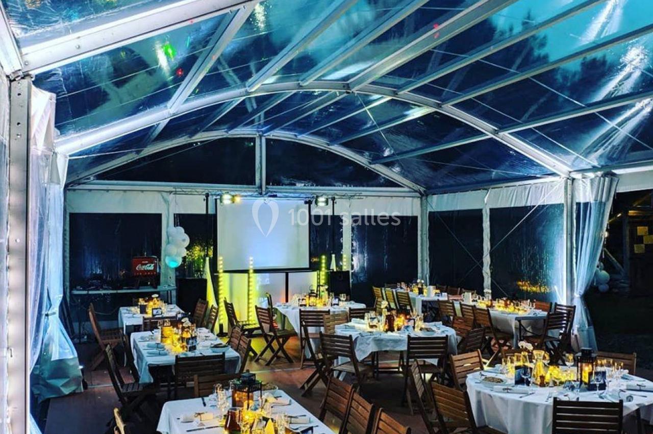 Salle sous chapiteau transparent avec tables dressées, chaises en bois, éclairage chaleureux et écran de projection.