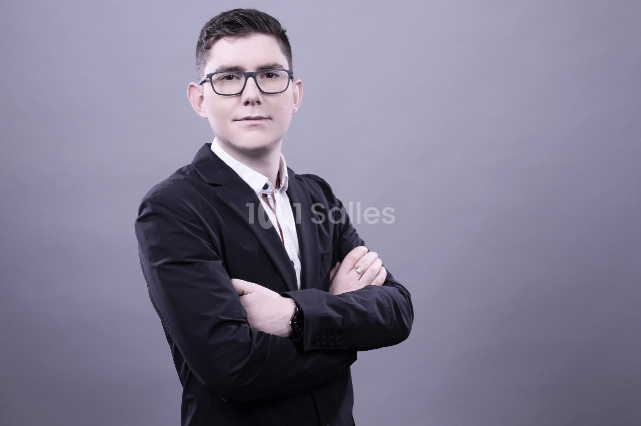 Homme en costume sombre, bras croisés, posant devant un fond gris uni.