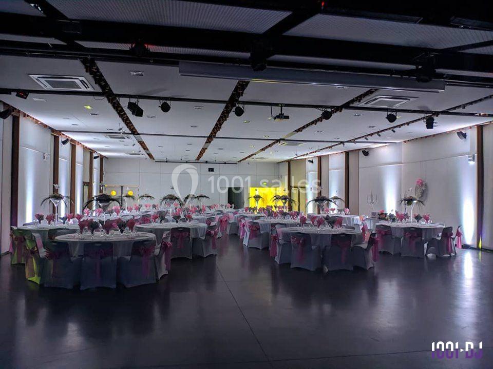 Salle de réception décorée avec des tables rondes dressées, nappes blanches et chaises ornées de rubans roses.