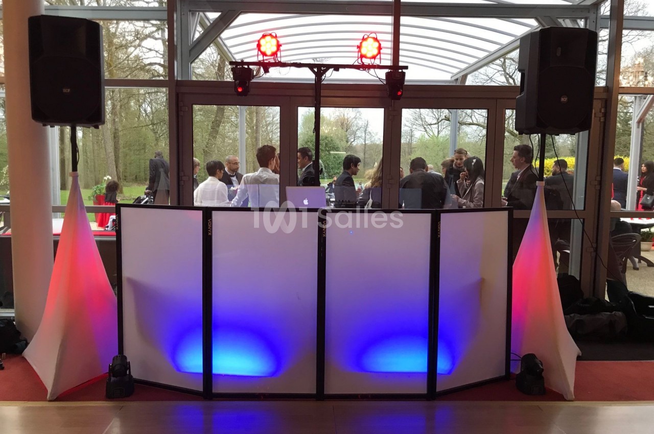 Console de DJ avec éclairage coloré devant une baie vitrée, des invités discutent en arrière-plan.