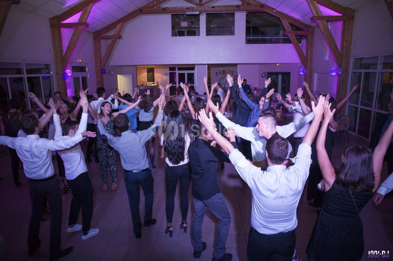 Des personnes dansent joyeusement dans une salle éclairée par des lumières violettes, avec une charpente en bois visible.