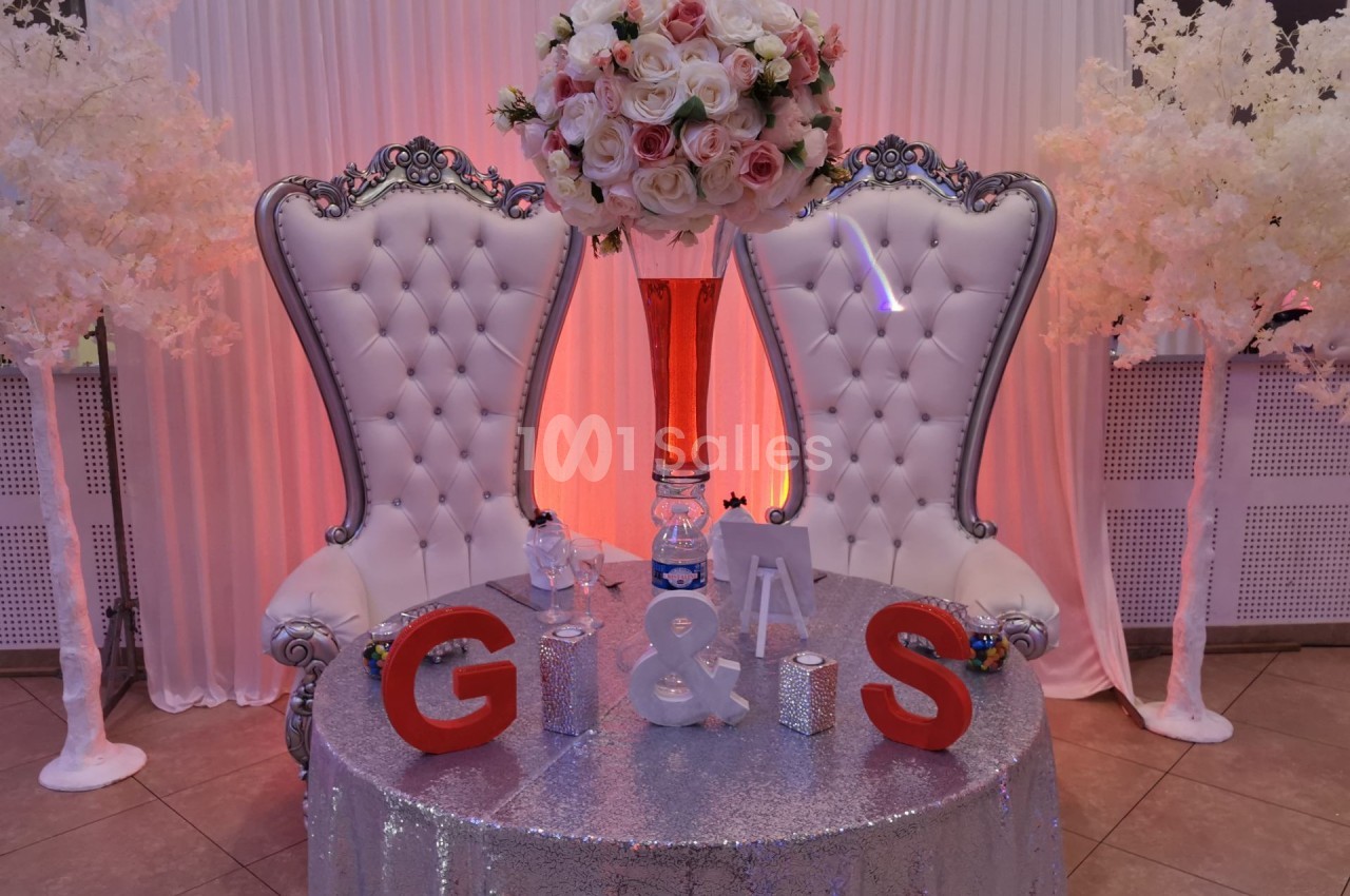 Deux fauteuils blancs capitonnés encadrent une table décorée avec des initiales, des fleurs et un vase rouge.