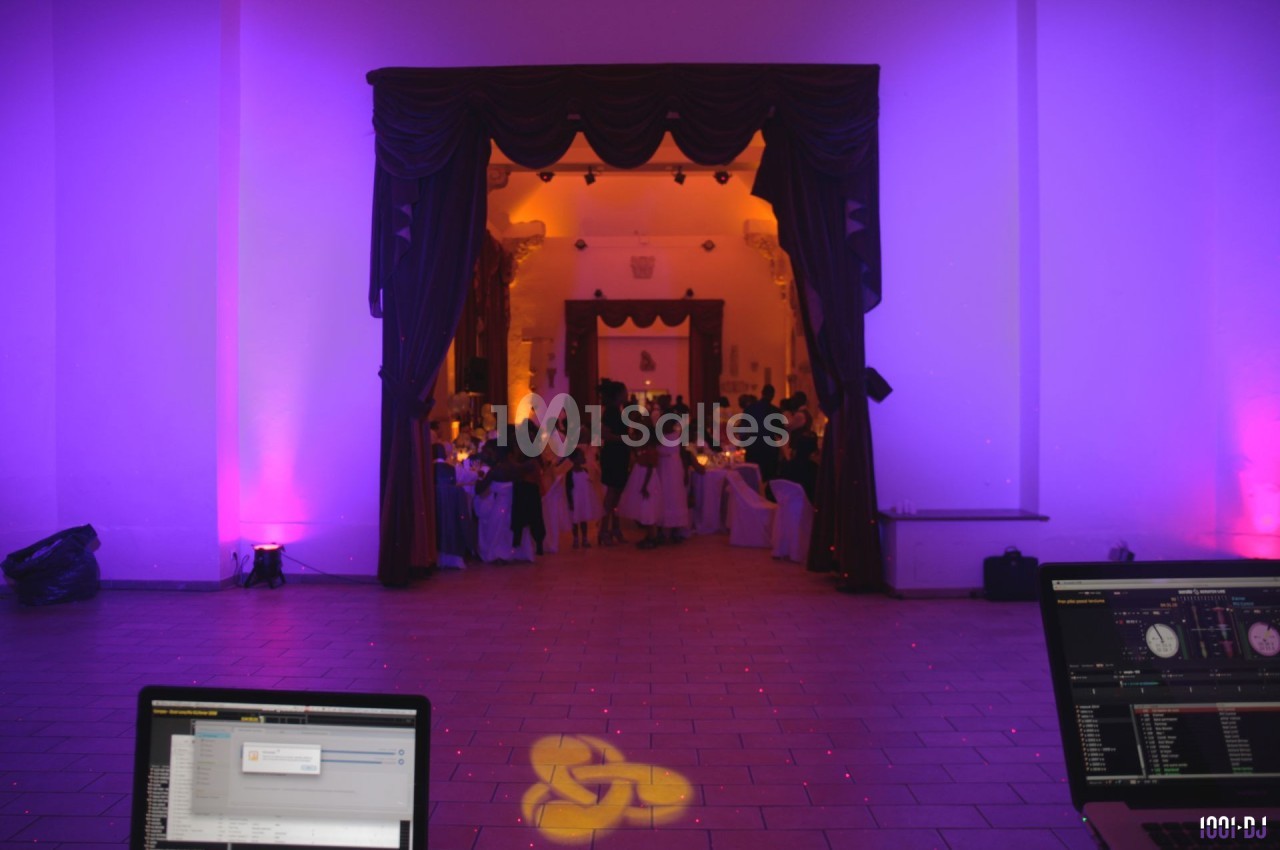 Salle de réception éclairée en violet et orange, avec des invités rassemblés au fond et des équipements de DJ au premier…
