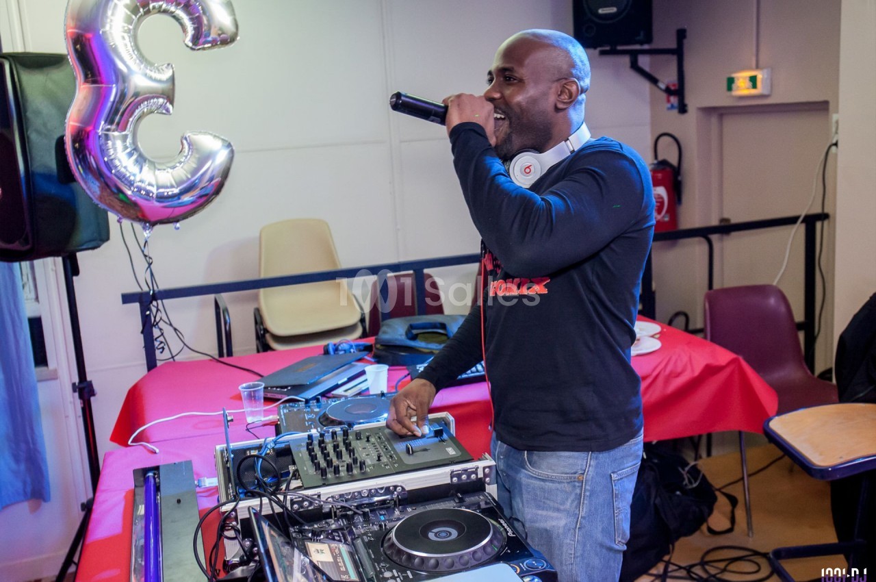 Un DJ mixe sur une table de mixage tout en chantant dans un micro, dans une salle décorée avec un ballon en forme de 3.