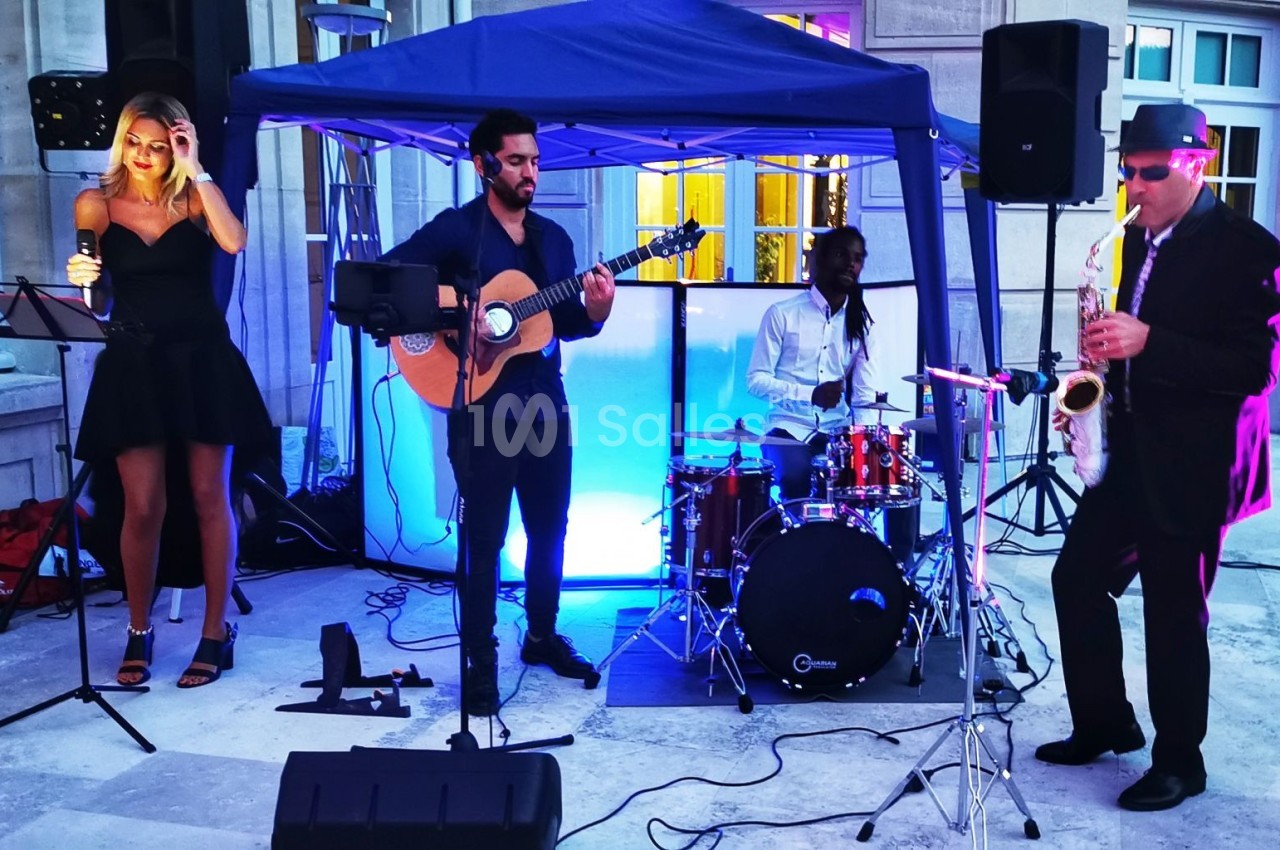 Un groupe de musiciens joue en extérieur sous une tente, avec une chanteuse, un guitariste, un batteur et un saxophoniste.