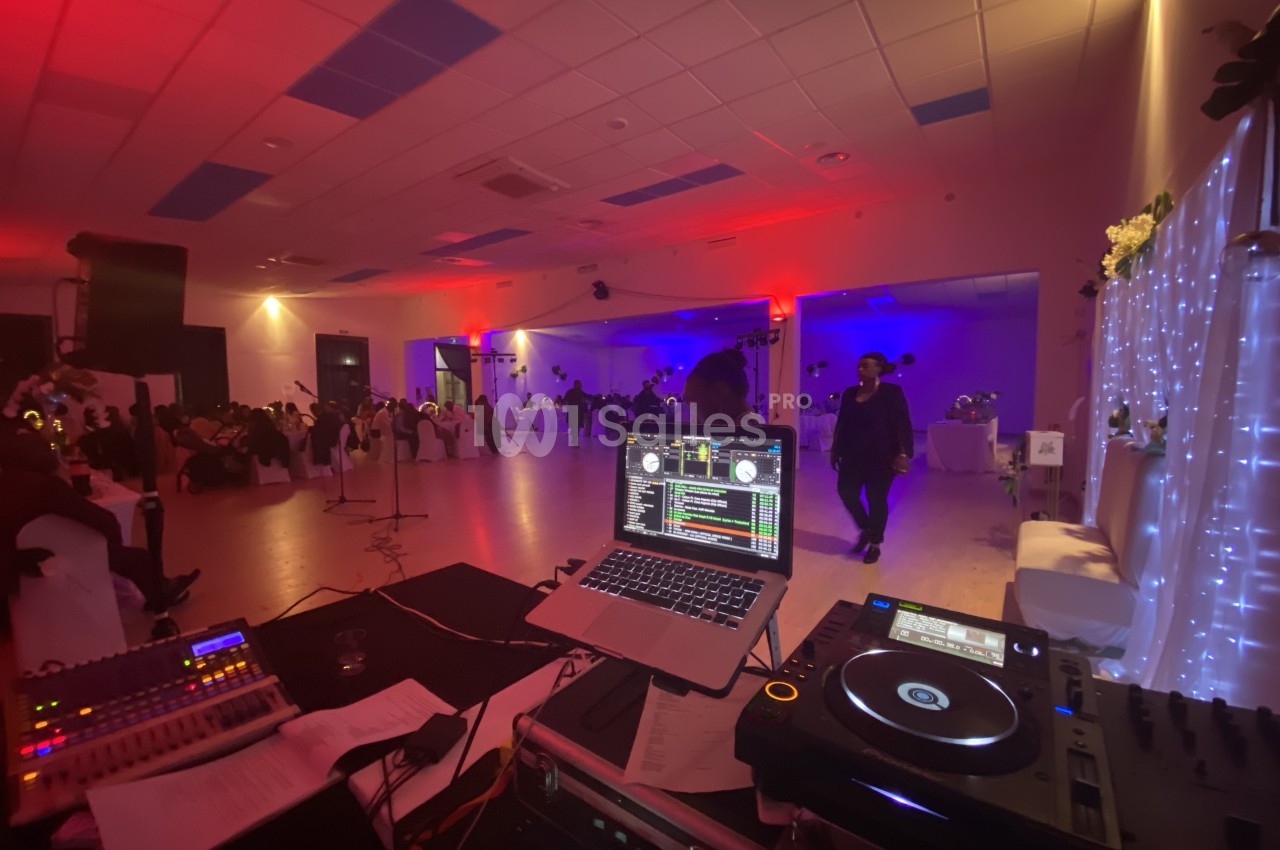 Salle de réception éclairée avec des lumières colorées, vue depuis une table de DJ équipée d'un ordinateur et de platines.