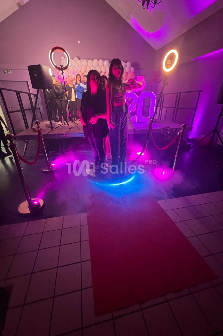 Deux personnes posent sur une plateforme lumineuse entourée de fumée, avec une scène décorée en arrière-plan.
