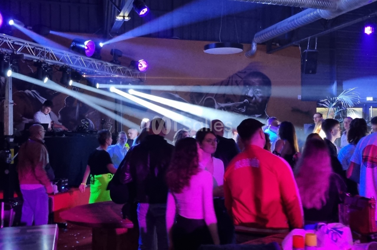 Des personnes dans une salle animée avec un DJ sur scène, éclairée par des projecteurs colorés.