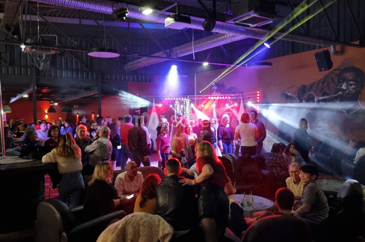 Salle animée avec des groupes de personnes discutant et dansant sous des jeux de lumière colorés.