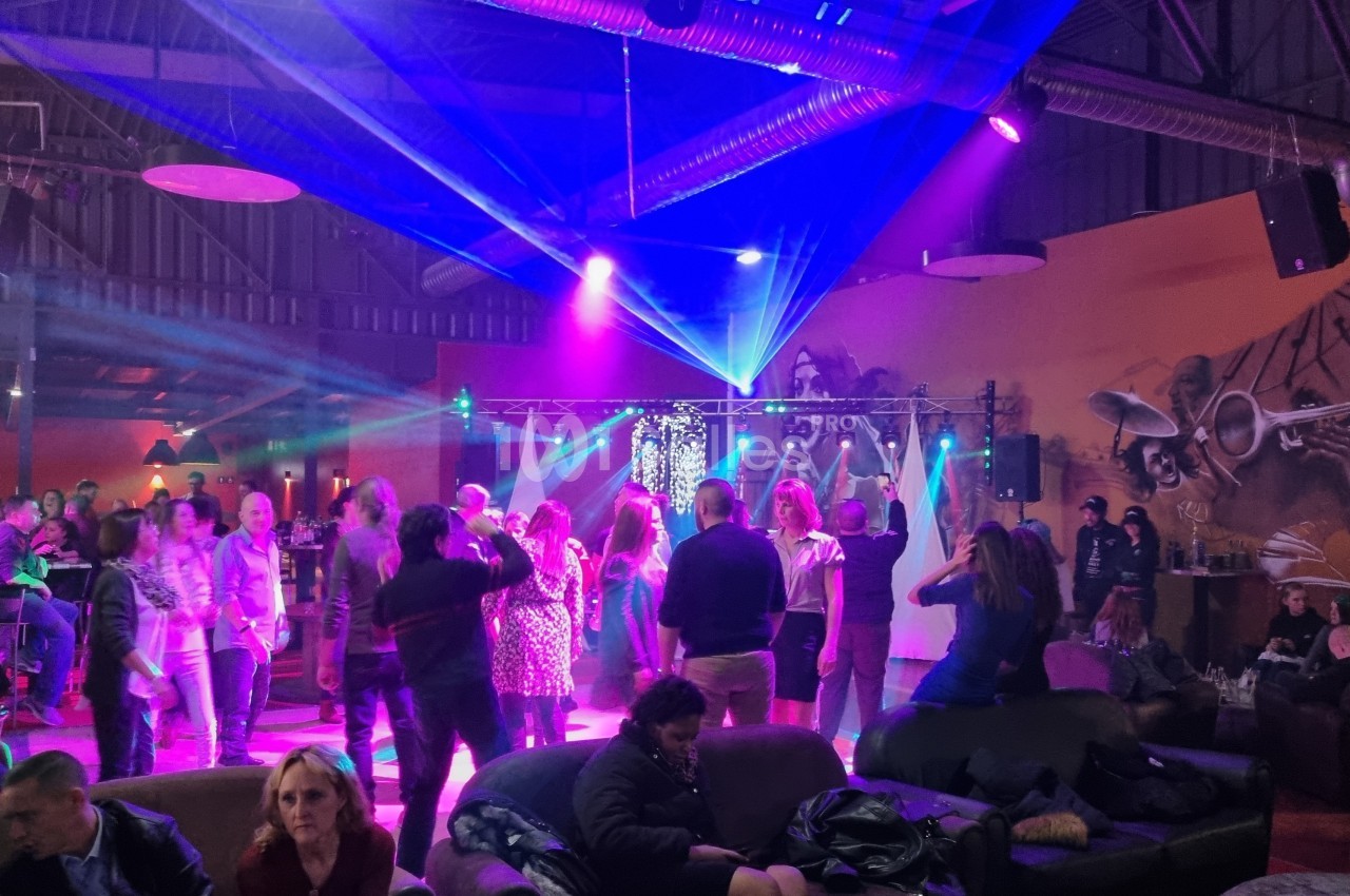 Des personnes dansent sous des jeux de lumière colorés dans une salle avec un décor mural musical et des spectateurs assis.