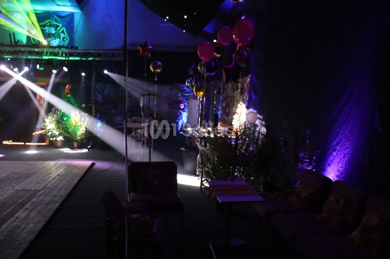 Salle sombre décorée avec des plantes, ballons et éclairages colorés, avec des sièges alignés sur le côté.