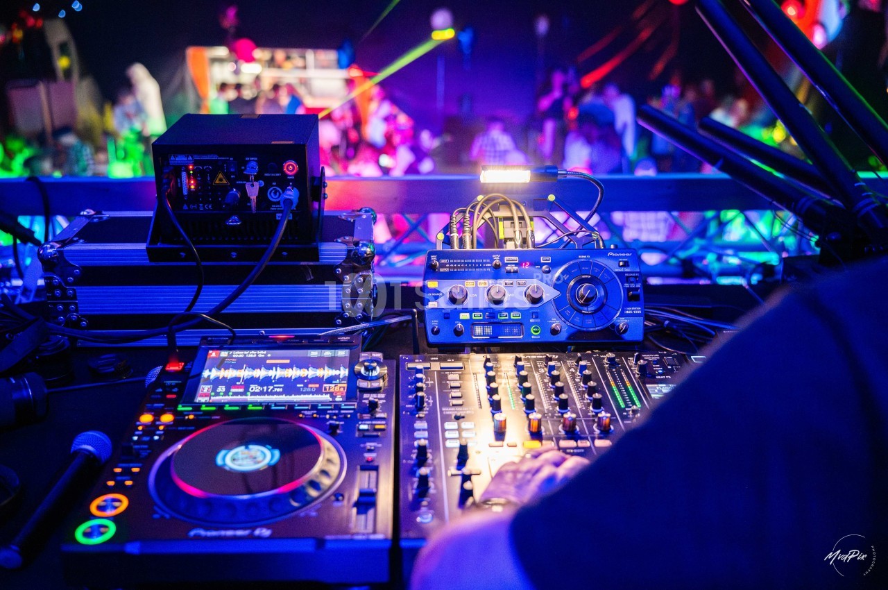 Vue rapprochée d'une table de mixage et d'équipements DJ, avec une piste de danse éclairée en arrière-plan.