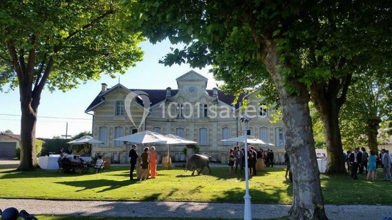 Personnes rassemblées dans un jardin devant une grande bâtisse en pierre, entourée d'arbres et de parasols.