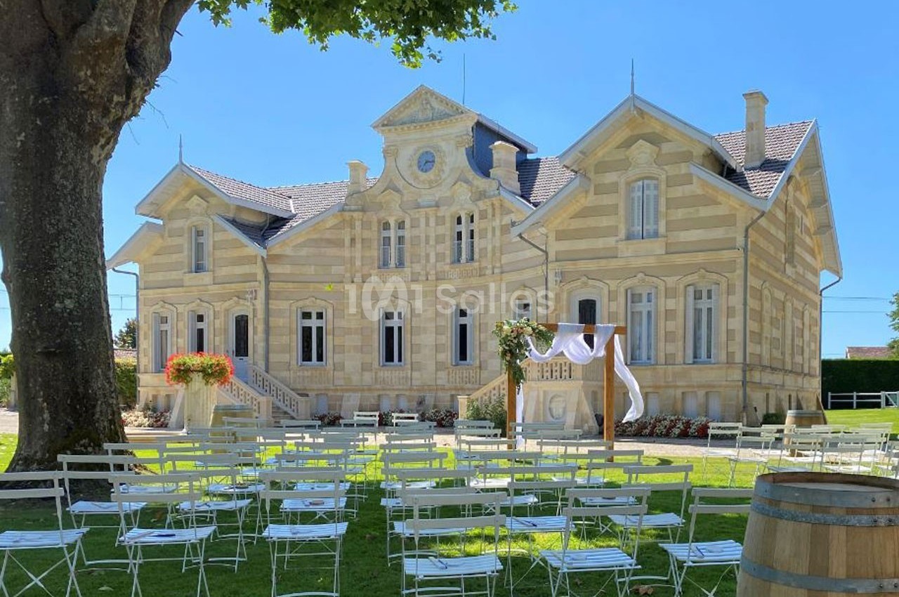 Façade d'une maison en pierre avec des chaises blanches disposées sur une pelouse pour un événement en extérieur.