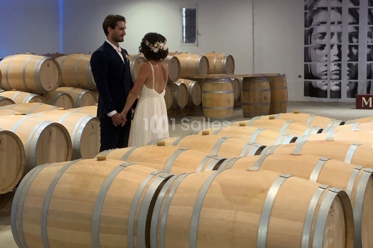 Un couple en tenue de mariage se tient la main dans une cave à vin entourée de fûts en bois.