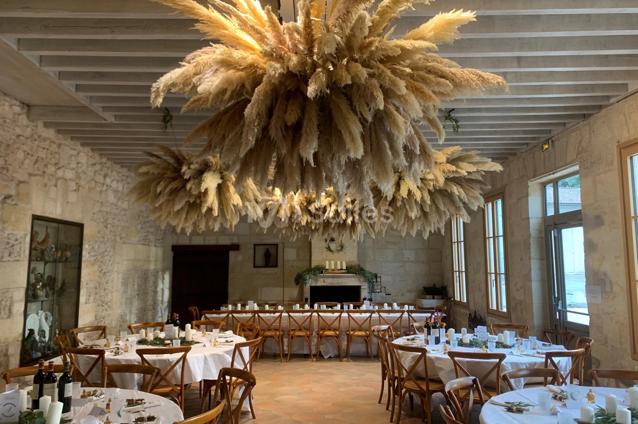 Salle de réception avec tables rondes dressées et grande suspension décorative en herbes de la pampa.