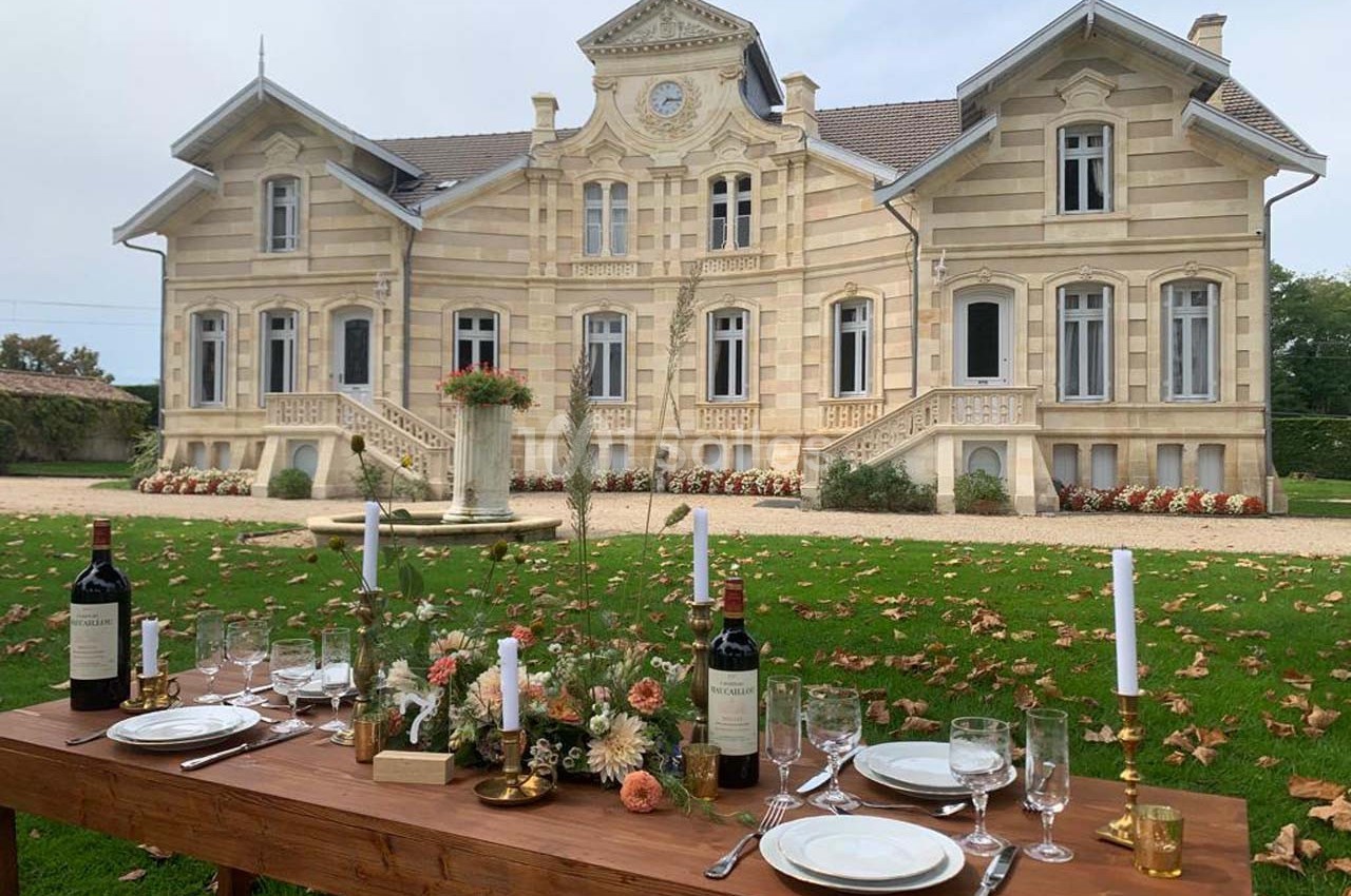 Table dressée avec vaisselle, bougies et fleurs devant un manoir en pierre entouré de pelouse et de feuilles d'automne.