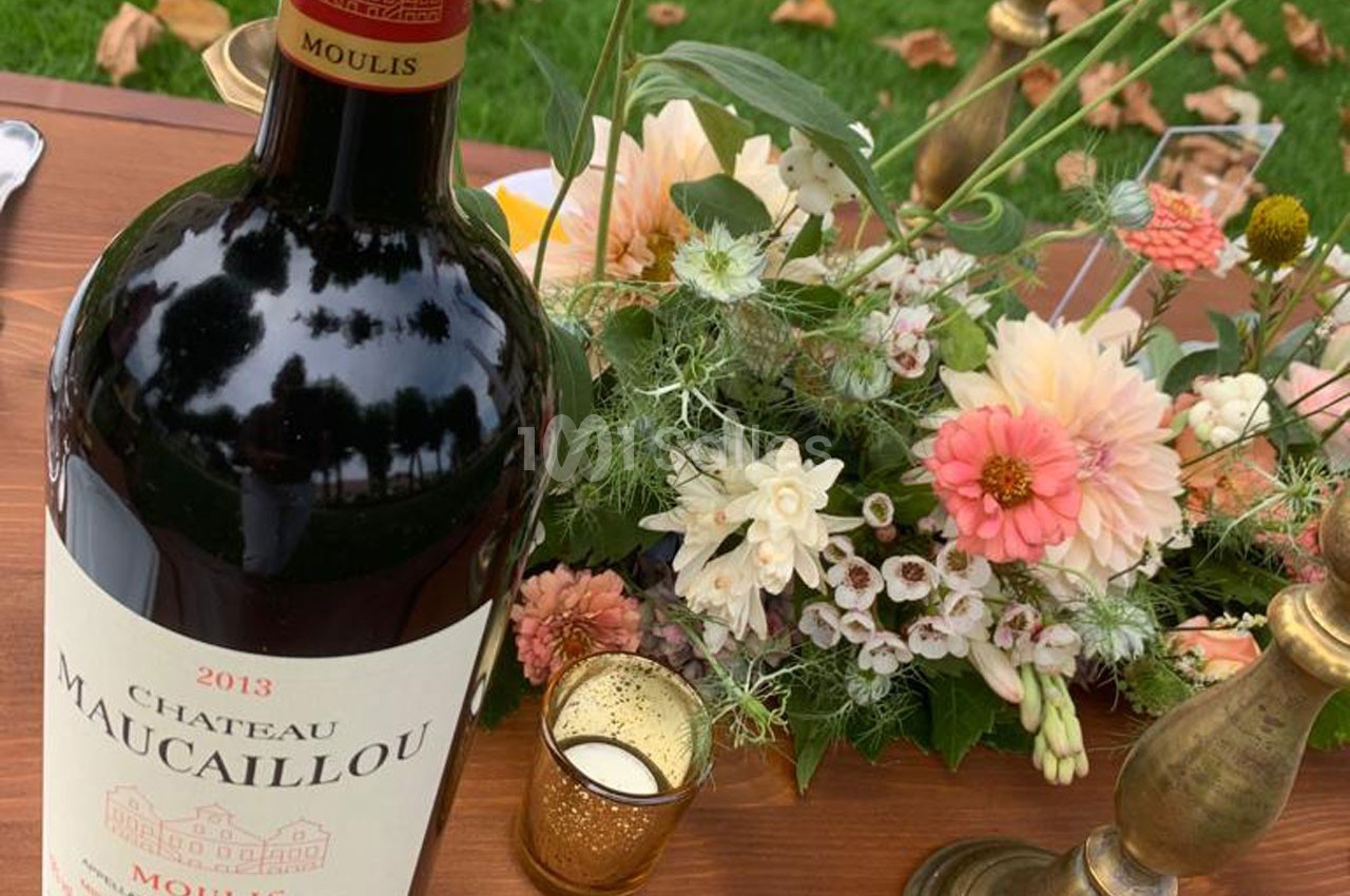 Bouteille de vin rouge Château Maucaillou 2013 posée sur une table avec un bouquet de fleurs et des bougeoirs.