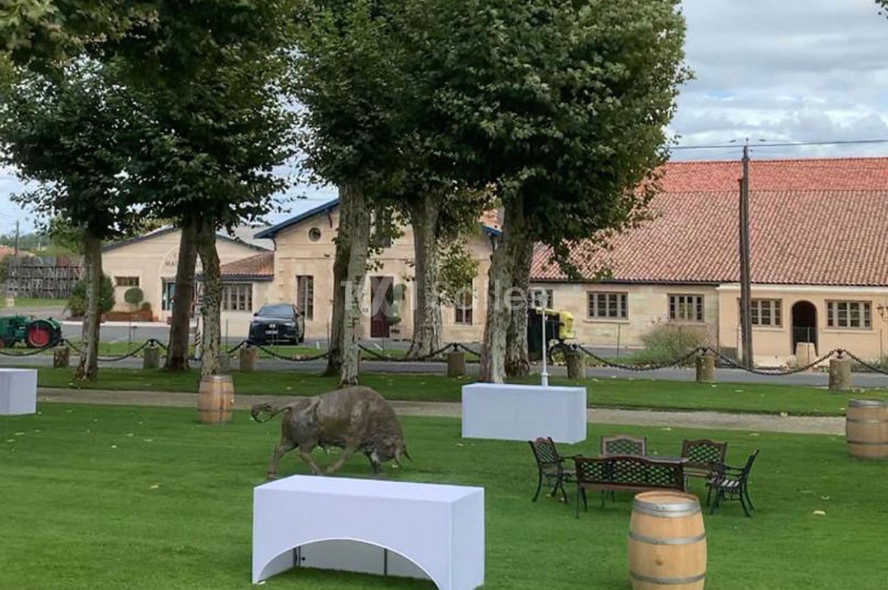 Statue de taureau sur une pelouse entourée de tonneaux, de tables et de chaises, avec des bâtiments en arrière-plan.